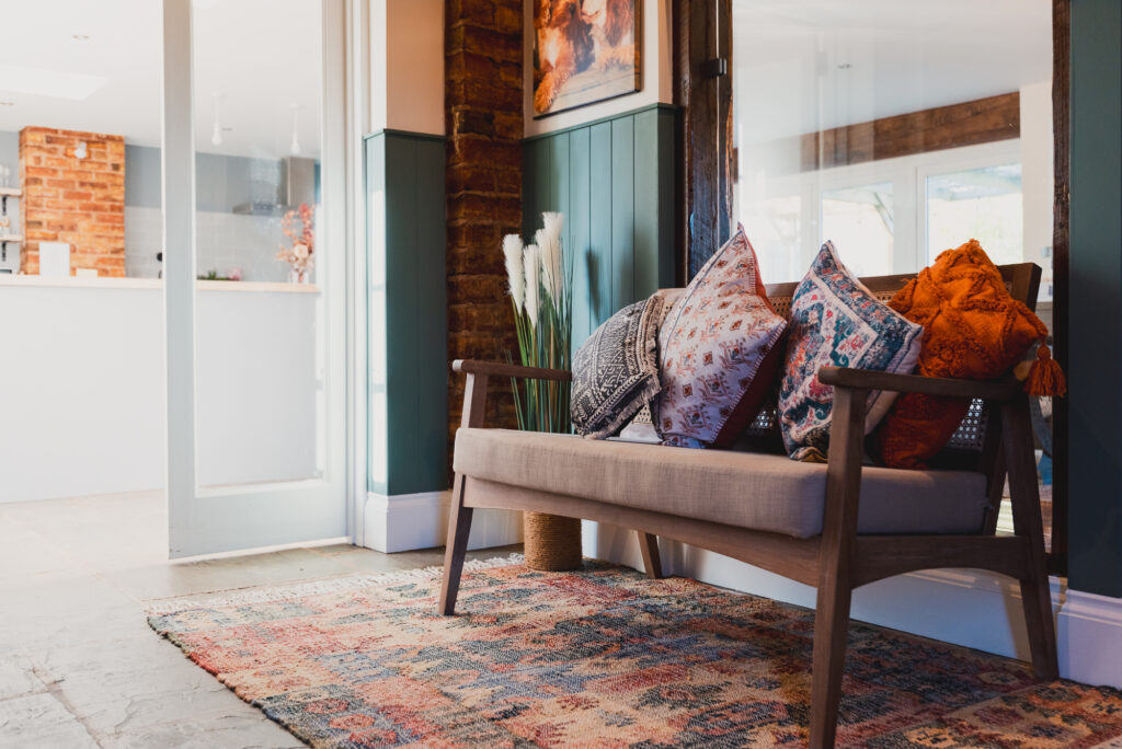 A wooden bench with colorful cushions sits on a patterned rug in a cozy, well-decorated room with exposed brick, green wainscoting, and a view into a modern white kitchen.