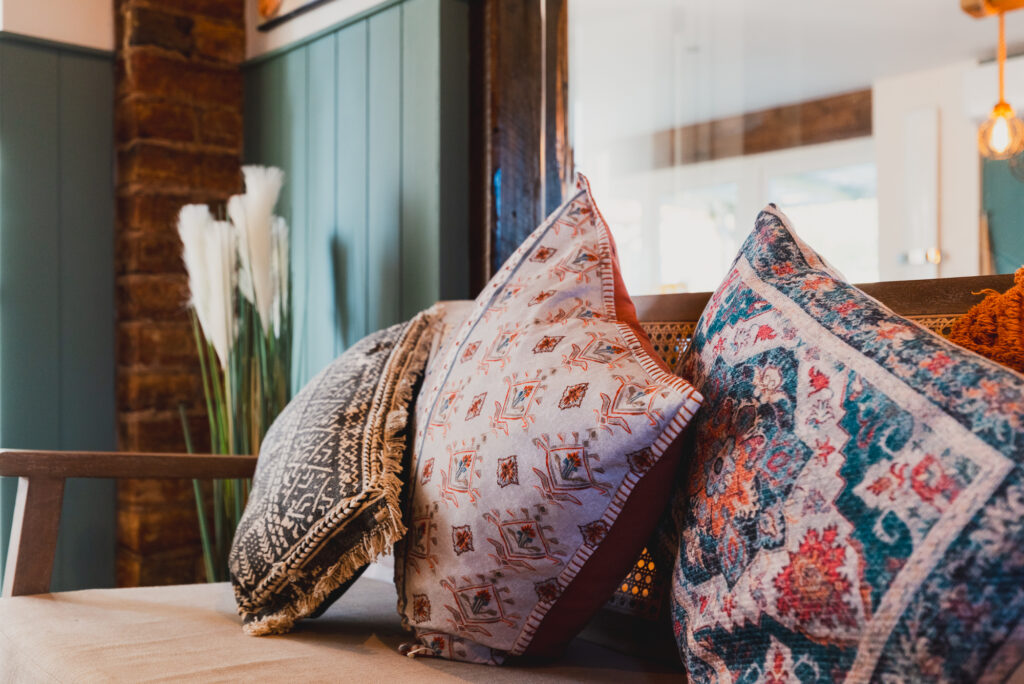 Decorative pillows rest on a beige cushioned bench in a cozy room with green paneling, brick accents, and tall white pampas grass in a vase.