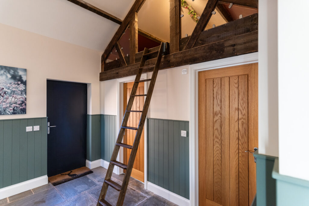 A wooden ladder leads to a loft above two wooden doors in a room with green wainscoting, grey tiled floor, a black door, and a floral wall picture.
