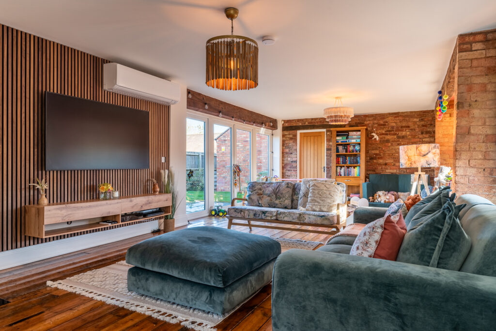 Television-mounted wooden feature wall in a modern living room, adorned with various cushioned sofas, chairs, and a wooden bookshelf, illuminated by ceiling lights and adjacent to floor-to-ceiling windows opening to a garden.